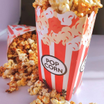 RED AND WHITE POPCORN CUP FILLED WITH BUTTERED POPCORN WITH A FEW KERNELS SCATTERED ON THE WOODEN TABLE.